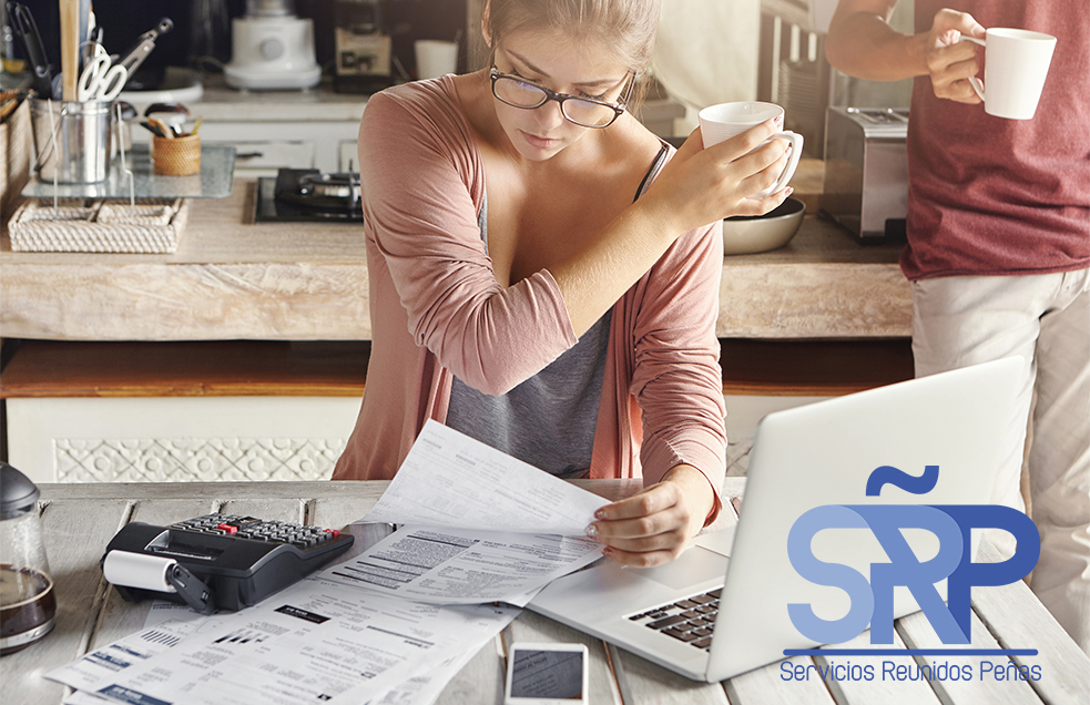 Concentrated woman dressed casually calculating bills, sitting at kitchen table with laptop, calculator, papers and mobile, holding white cup and passing it to her husband, who is standing behind her